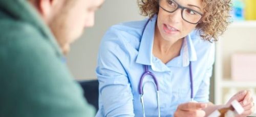 Vrouwelijke huisarts in gesprek met een cliënt die naar een buisje pillen kijkt.